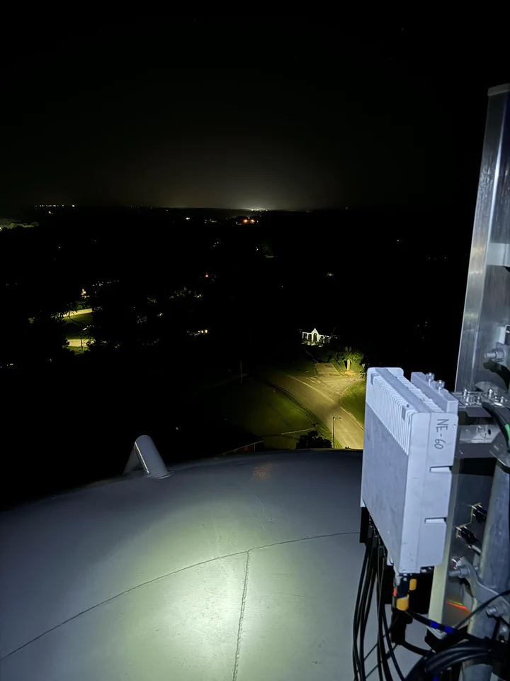Vertical Axis tower-top install on an Alabama Lightwave water tower at night, with equipment mounted on the tank roof and city lights on the horizon