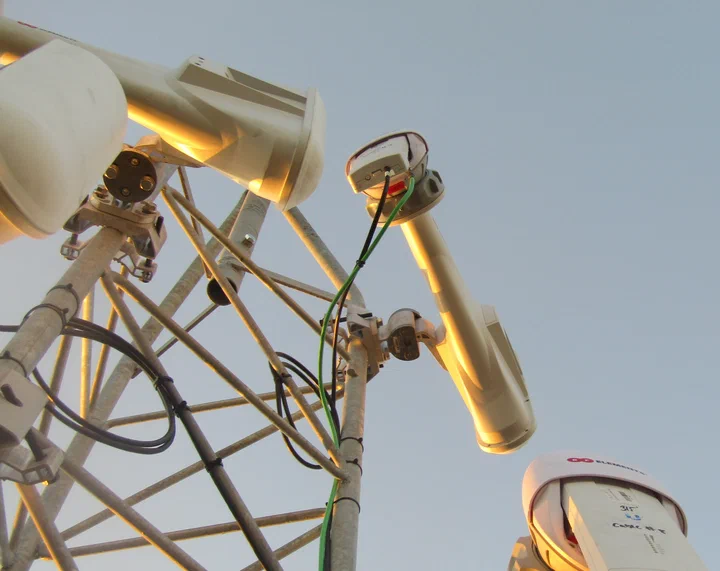 Upward view of an RF Elements horn antenna and small radio mounted on a red lattice tower leg against a blue sky streaked with cirrus clouds
