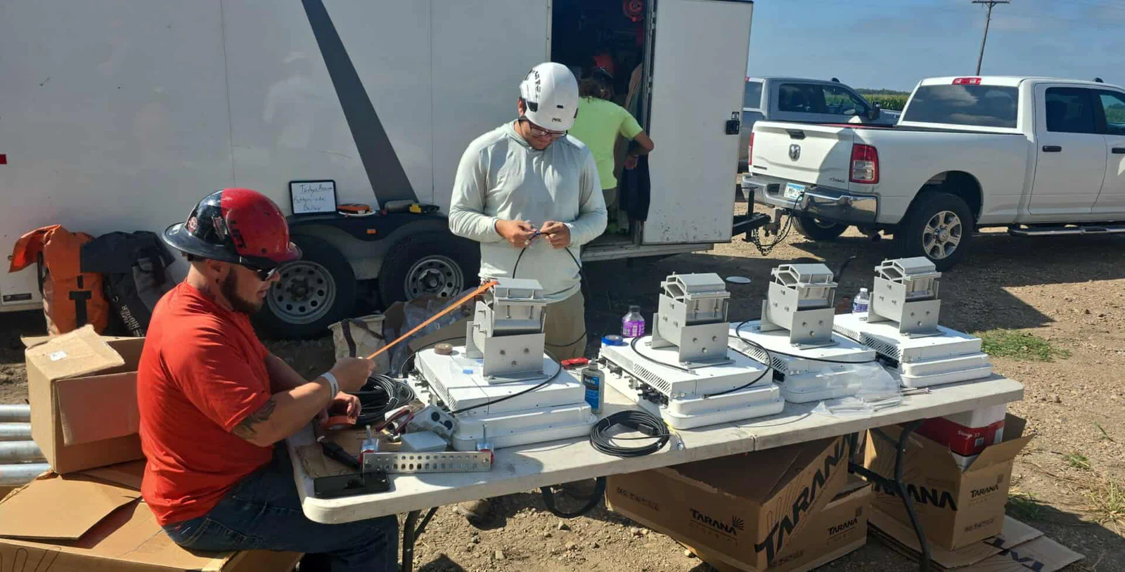 Vertical Axis crew pre-testing a row of Tarana radios on a folding table at the site, enclosed gear trailer and pickup staged behind them, rural sunlit terrain in the background. Ground-level staging before anything climbs the tower.