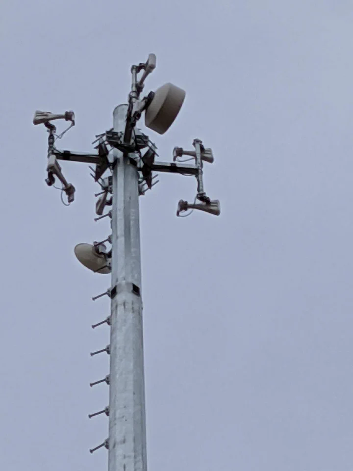 Close-up of the crown of a completed direct embedment monopole showing backhaul dish, sector antennas, and step-bolts running down the pole