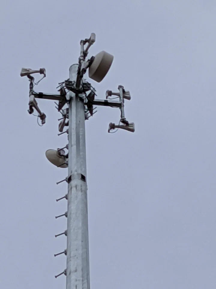 Close-up of the crown of a completed direct embedment monopole showing backhaul dish, sector antennas, and step-bolts running down the pole