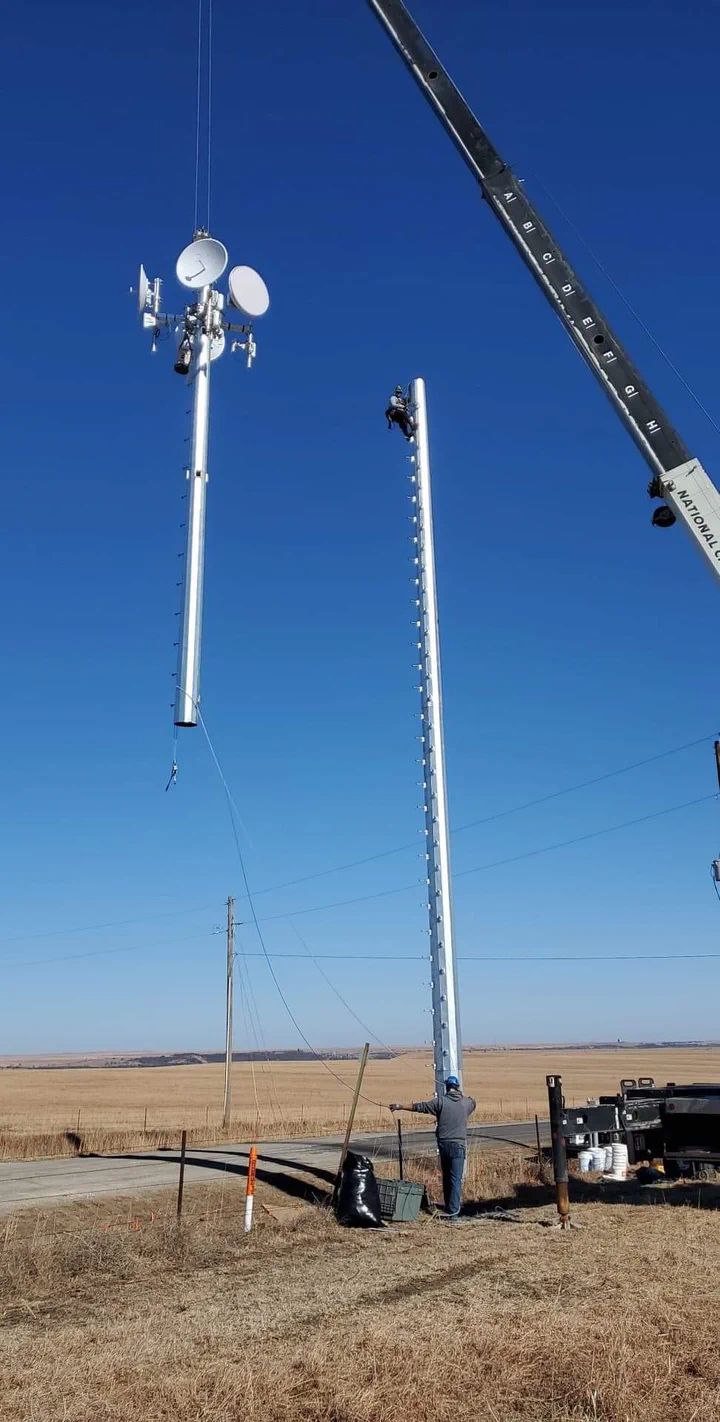 Crane flying two direct embedment monopole sections into position on a rural prairie site, one section already topped with backhaul dishes and sector antennas while a climber rigs on the second