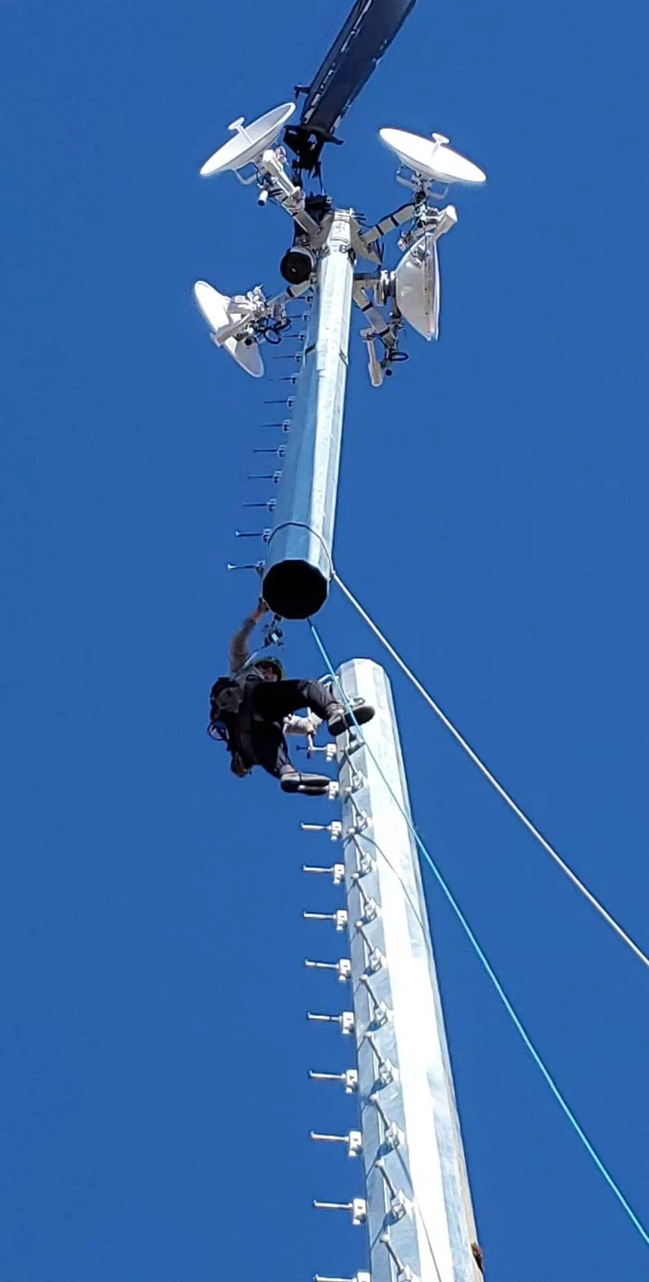 Vertical Axis climber rigged on a direct embedment monopole during install, crane lowering a section overhead and the full antenna array visible at the crown