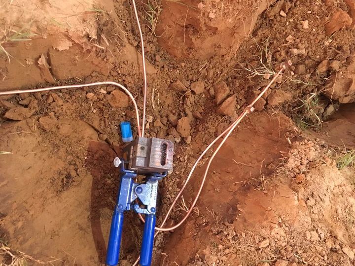 Cadweld T-splice mold clamped in blue handle tooling over three bare copper ground conductors in the open trench, ready for the exothermic powder charge that will fuse the bond