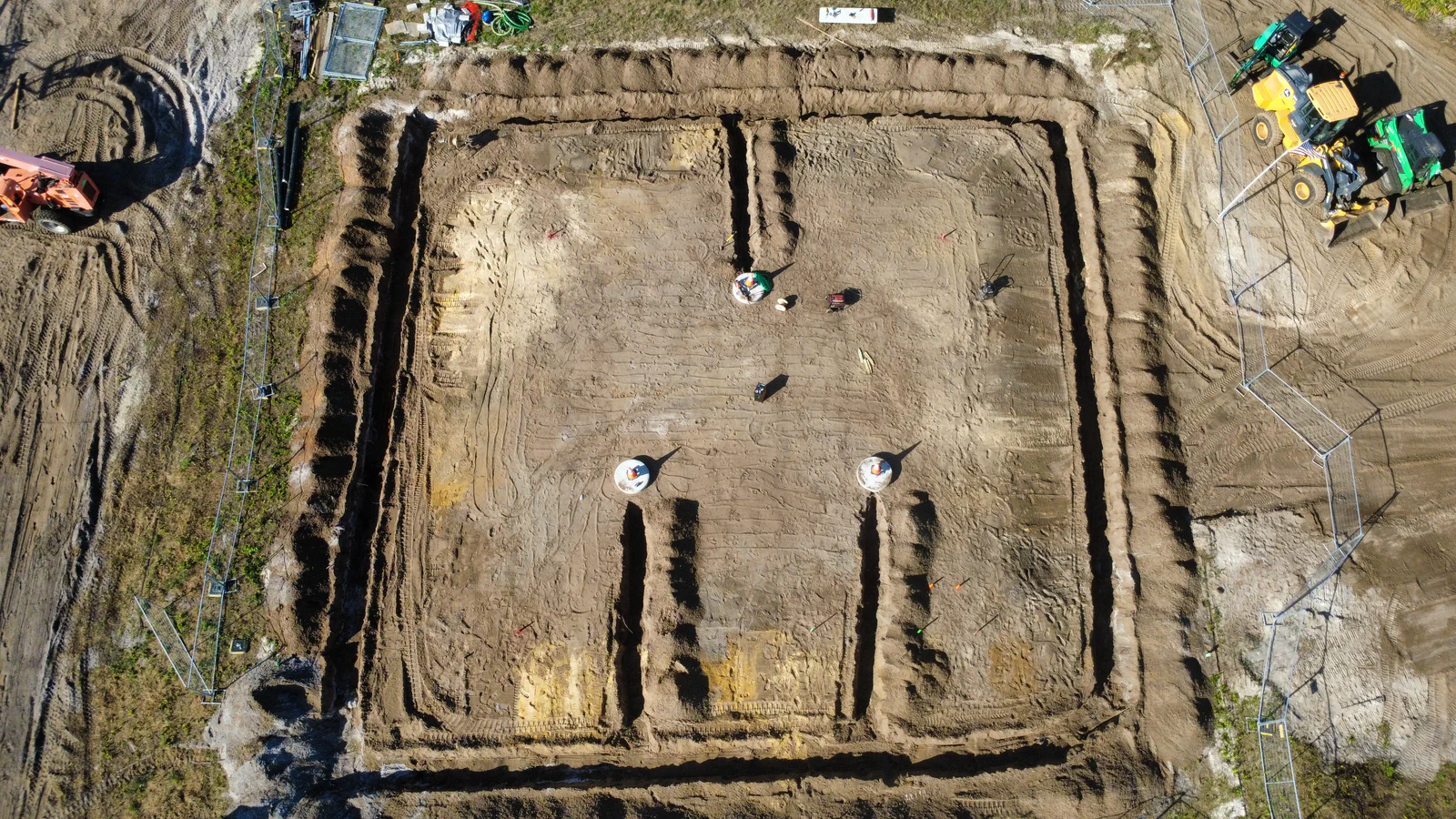 Drone view straight down on a new tower pad with the ground ring trenched around the perimeter, foundation piers poured in the center, and the crew laying in tinned copper conductor before backfill