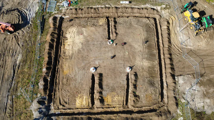 Drone overhead of the full perimeter ground-ring trench around a new tower pad, foundation piers centered, conductor and rods staged for cadweld bonding