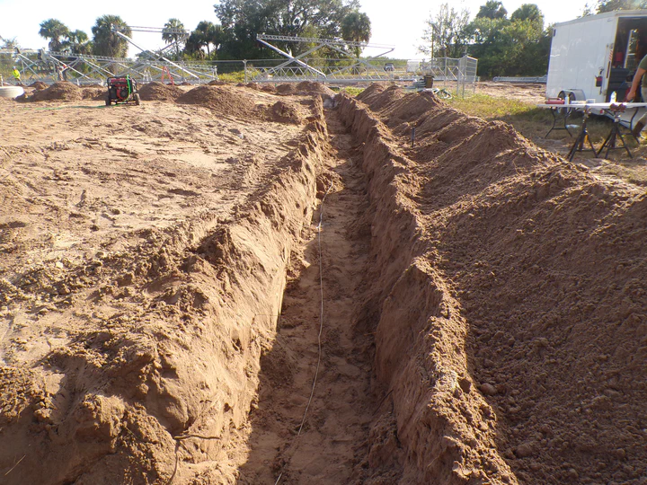 Open ground-ring trench running the length of the pad with bare tinned copper conductor laid in the bottom, tower steel and crew trailer visible at the far end of the cut