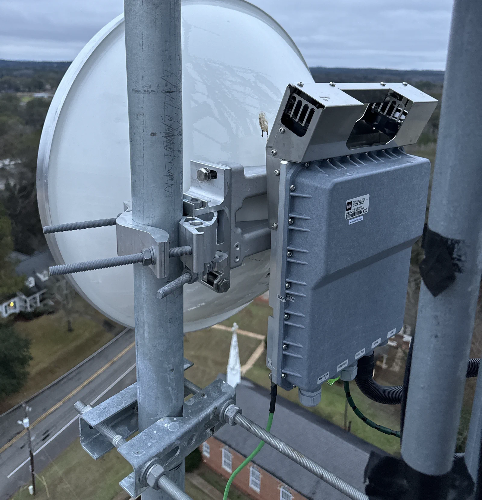 Close-up of a carrier-grade SAF Tehnika microwave ODU paired with a white high-performance dish on a Vertical Axis tower leg, town skyline and road visible below