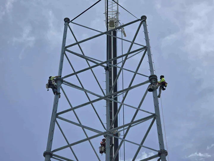 Two Vertical Axis climbers rigged at the crown of a new self-supporting lattice tower working the final top-section connections with a third climber staged on the monopole core below