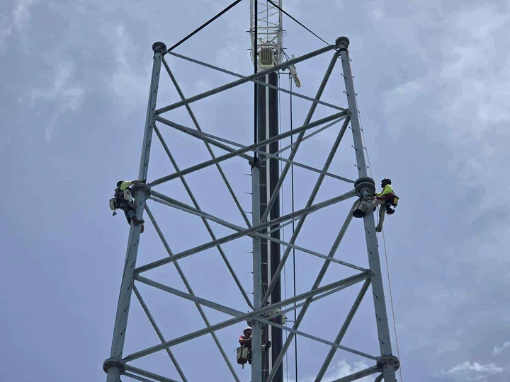 Two Vertical Axis climbers rigged at the crown of a new self-supporting lattice tower working the final top-section connections with a third climber staged on the monopole core below