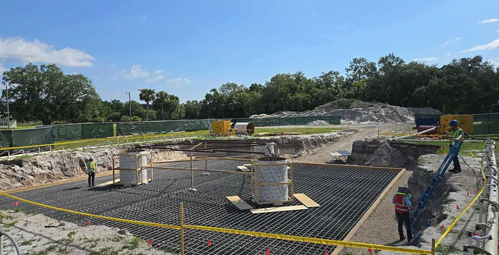 Daytime shot of a new tower pad with two anchor-bolt template piers formed up, rebar mat tied across the excavation, and Vertical Axis crew staking layout and walking the grade