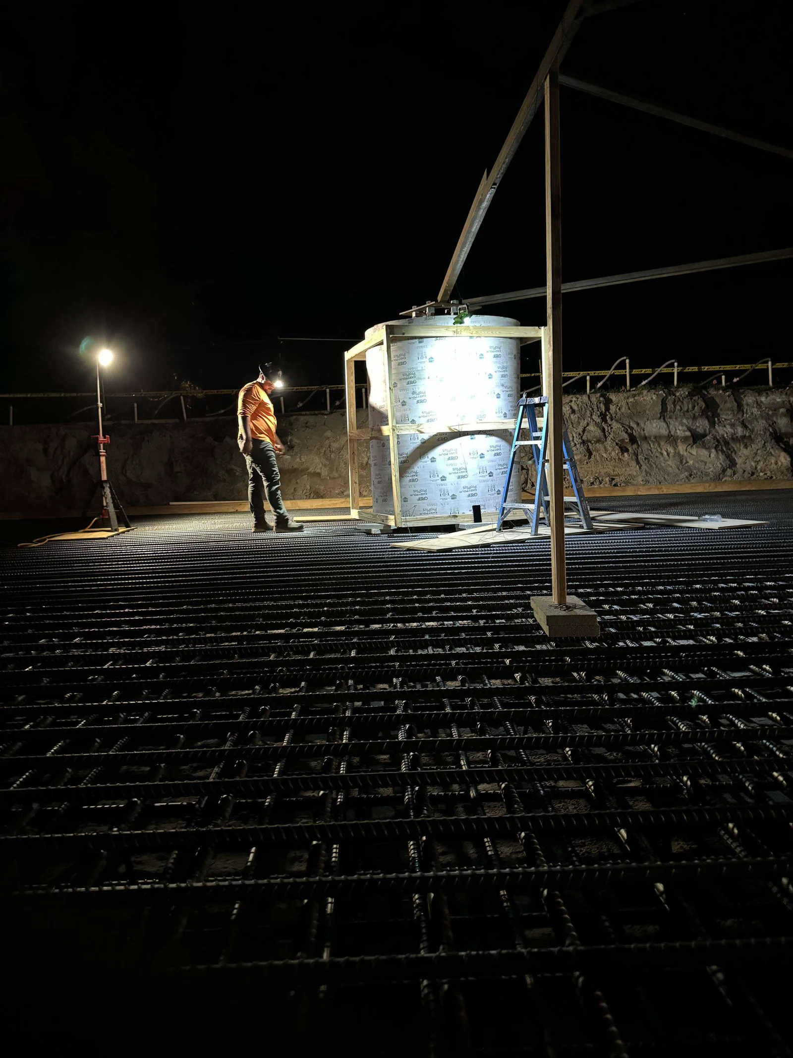 Night pour prep at a new tower pad: crew member working under portable work lights next to a lit anchor-bolt template cage set over a freshly tied rebar mat ready for concrete