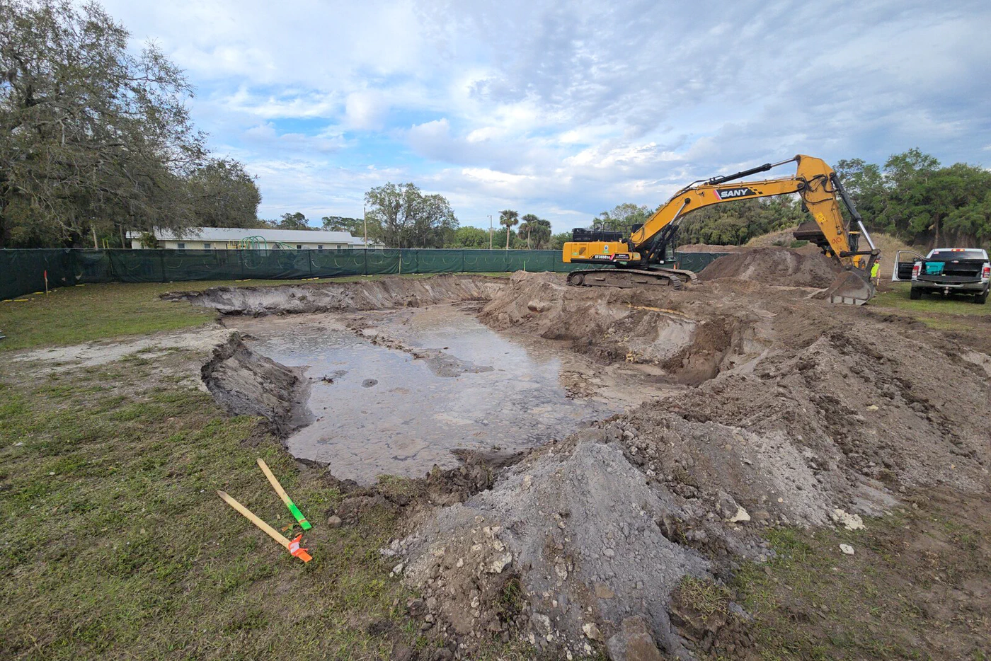 Excavated tower foundation pit with standing water, spoil piles staged off-pad, and a track hoe on the far side