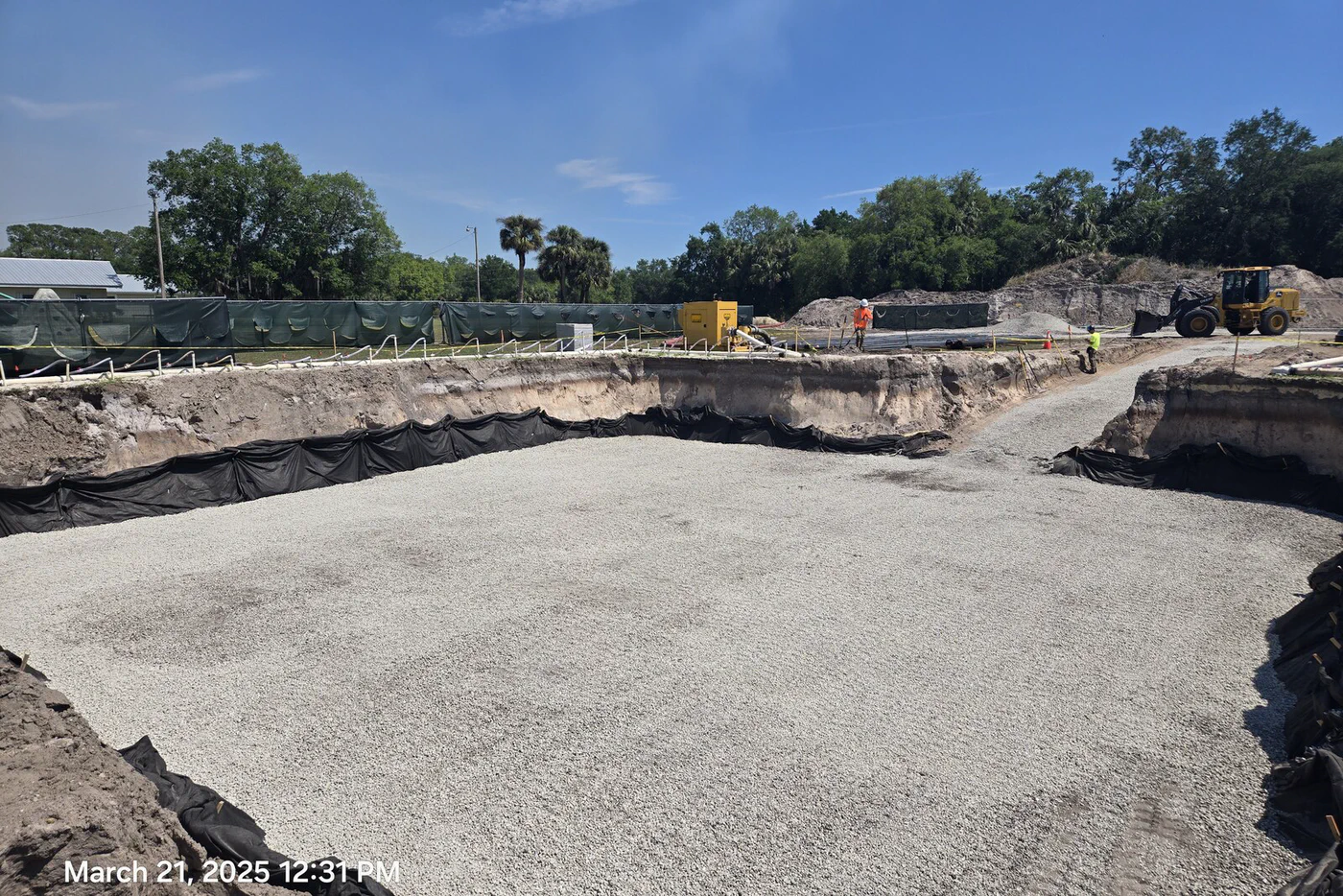 Compacted crushed-stone base inside the formed foundation pit, geotextile fabric lining the walls, a mini excavator and operator on the far side