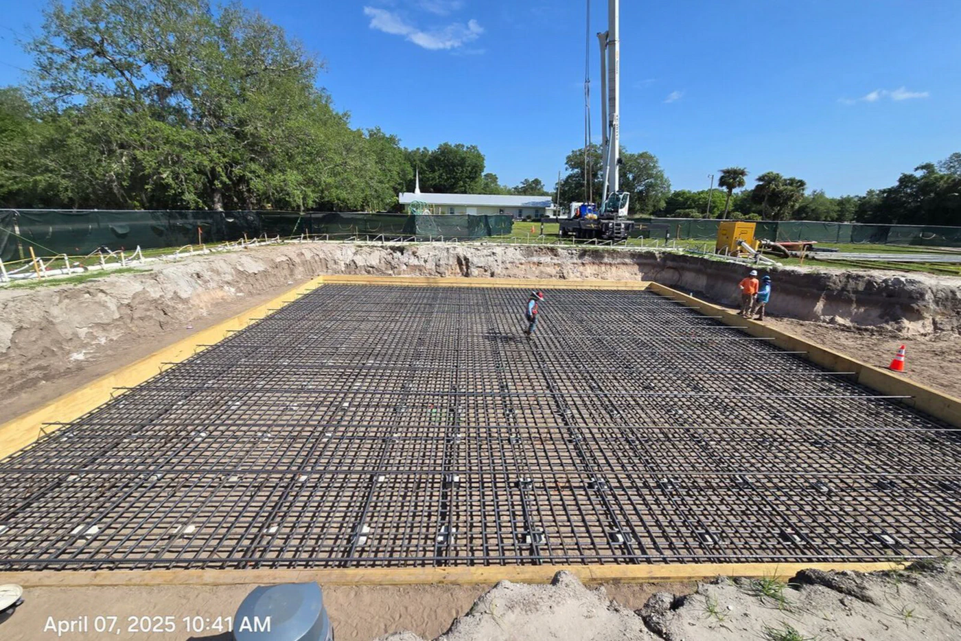 Tower foundation with a full rebar mat laid out on the compacted base, a drilled-pier rig staged on the far side, and a crew member inside the rebar grid