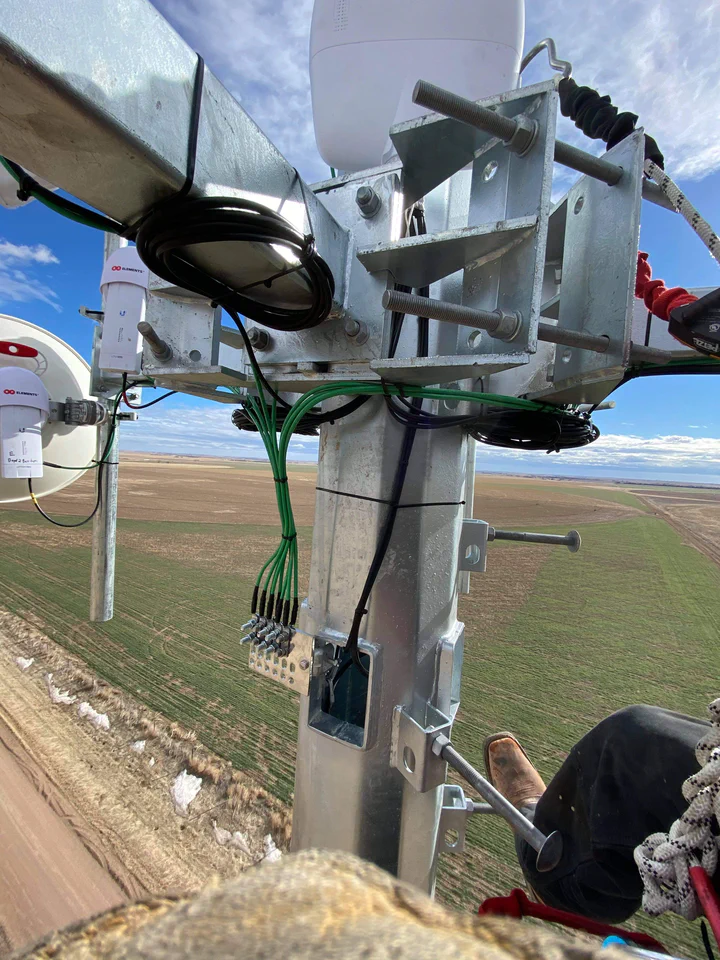 Climber's view looking down at a sector-antenna mount: dressed RF jumpers, clipped Cat6 runs, green bonding conductors tied to the bracket, and the rural horizon visible hundreds of feet below