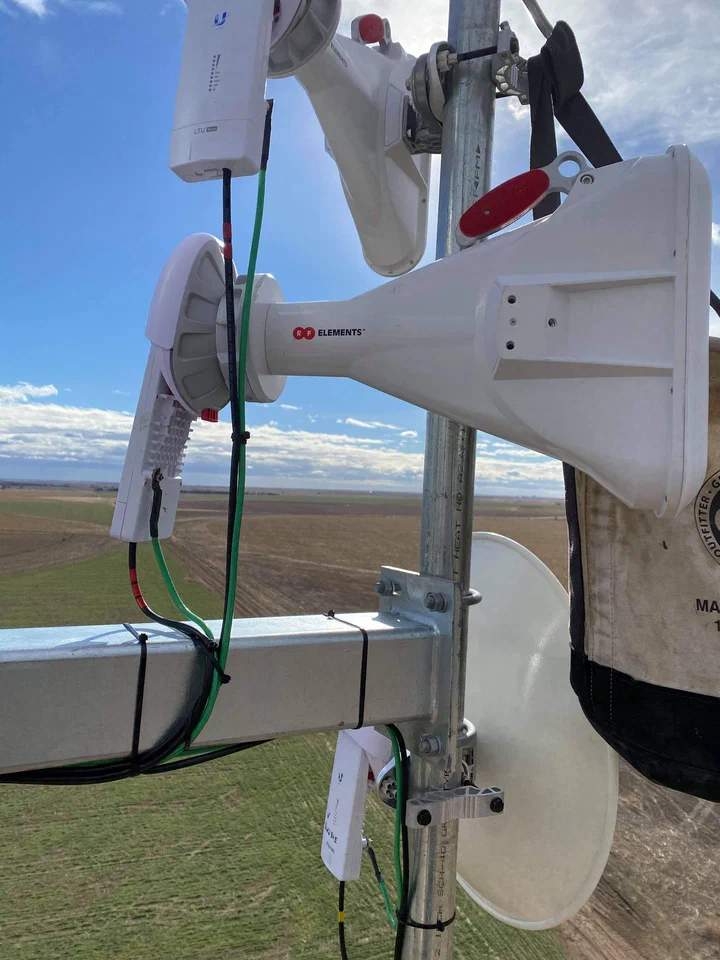Tight tower-top install: an RF Elements horn and a Ubiquiti backhaul paired on a single mount pipe with dressed jumpers, green bonding conductors, and a climber's tool pouch hanging off the bracket