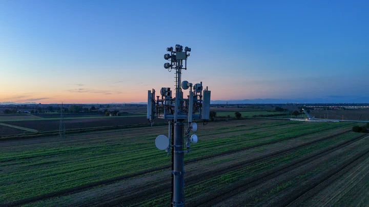 Dusk aerial of a deployed multi-sector tower standing in open farm fields at sunset, the built output of a Vertical Axis site-design package run from coverage brief to commissioned site