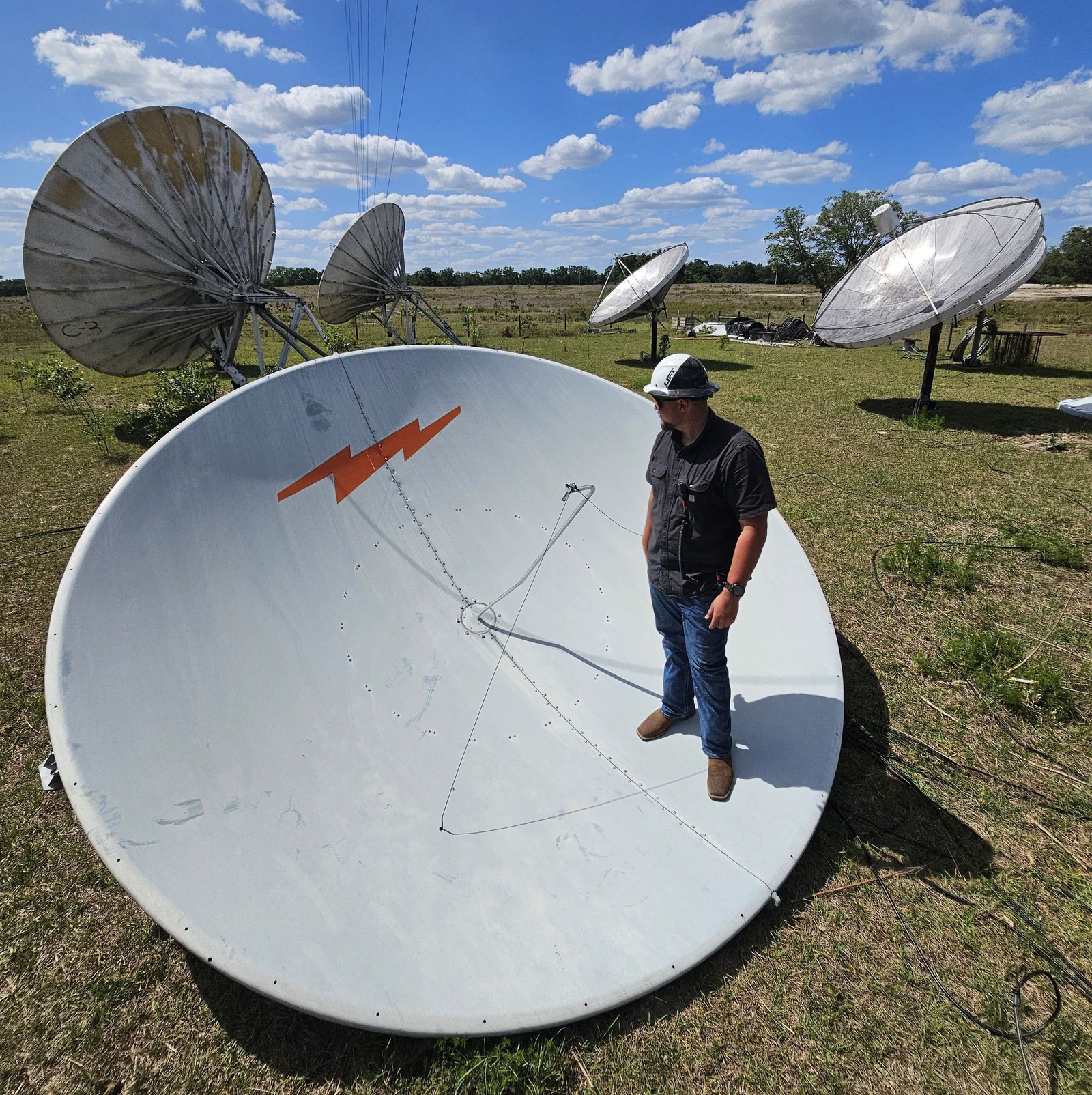 Vertical Axis crew member in hard hat standing on a large decommissioned satellite dish laid flat in an open field, four other dishes down on the grass behind him, rural landscape under summer cumulus clouds.
