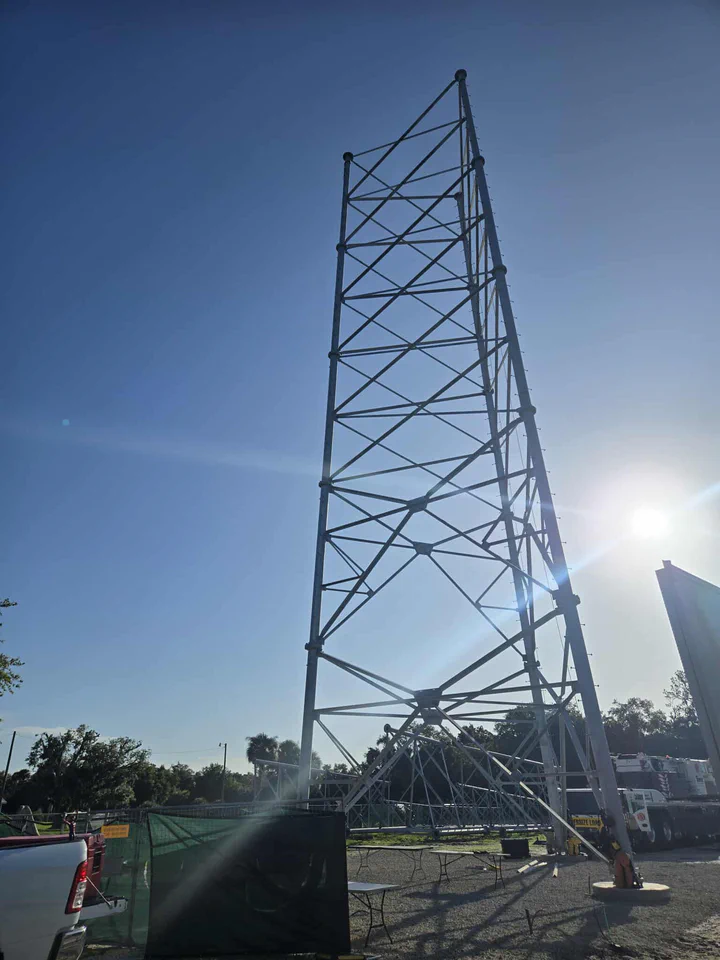 Dramatic low-angle view of a completed self-supporting lattice tower silhouetted against the sun with the mobile crane and oversize-load trailer still staged at the base