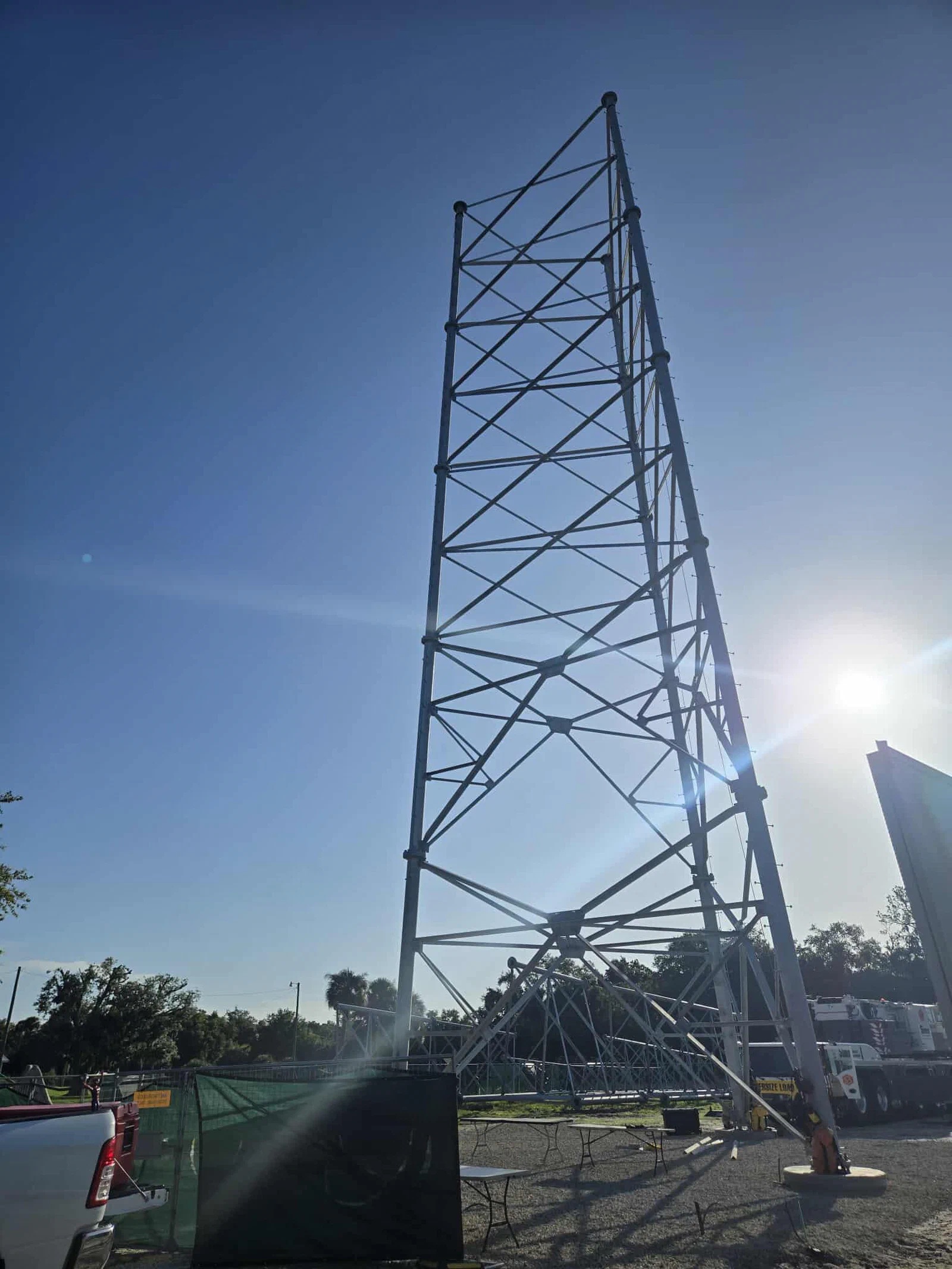 Lower sections of a self-supporting lattice tower standing on pier foundations mid-stack, with the crane and oversize-load trailer staged on the compound