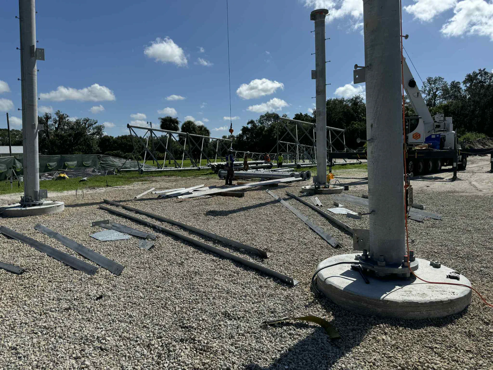 Self-supporting tower base set on anchor-bolt piers with steel lattice members staged on the pad and a mobile crane rigged in the background ready for section flies